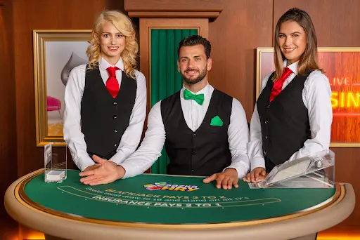 Živí krupiéři u stolu pro blackjack v online kasinu. Tři krupiéři v živém kasinu stojí za stolem pro blackjack a usmívají se do kamery.